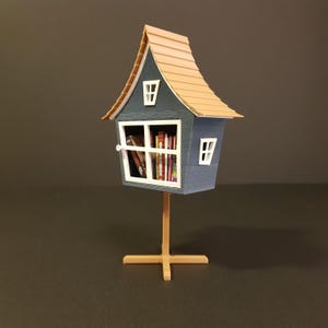 Peut inclure: Un stand d'échange de livres miniature en forme de maison bleu et marron avec une fenêtre et une étagère pleine de livres. Le stand est sur une base en bois avec un support en forme de croix.