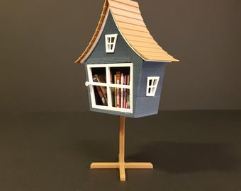 Może przedstawiać: Miniaturowy stojak na wymianę książek w kształcie domu w kolorze niebieskim i brązowym z oknem i półką pełną książek. Stojak stoi na drewnianej podstawie z krzyżowym wspornikiem.