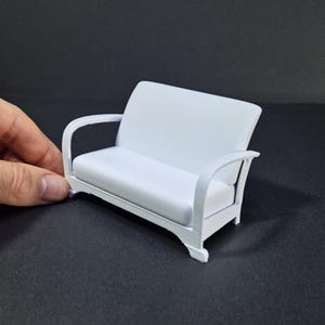 May include: A small, white miniature sofa with curved armrests and a cushioned seat and back. The dollhouse-sized furniture piece is made of a solid, opaque material and is designed to resemble a classic loveseat.