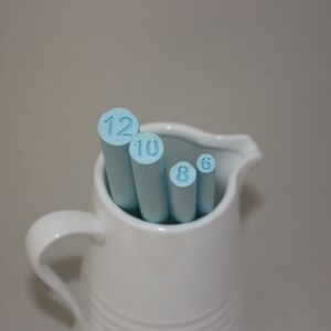 May include: A white ceramic pitcher with a handle, holding four blue painted wooden dowels with the numbers 12, 10, 8, and 6 printed on them.