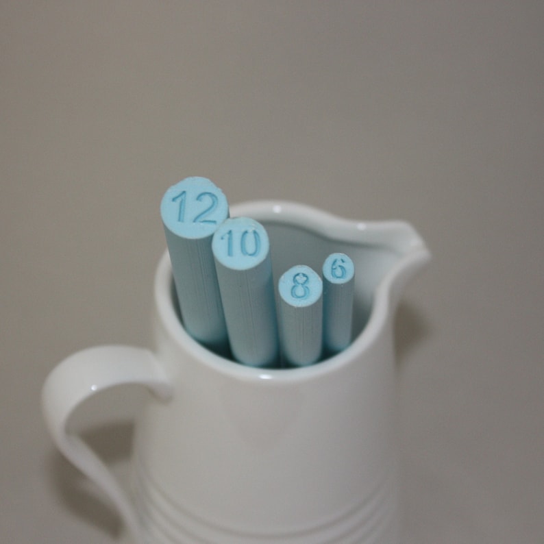 May include: A white ceramic pitcher with a handle, holding four blue painted wooden dowels with the numbers 12, 10, 8, and 6 printed on them.