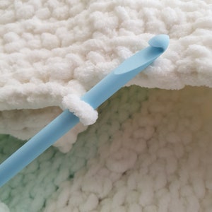 May include: A close-up of a light blue crochet hook resting on a white and light green crocheted fabric.