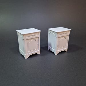 May include: Two white miniature nightstands with a drawer and a decorative door. The nightstands have a classic design with curved legs and ornate detailing. The nightstands are made of a solid material and are suitable for a dollhouse or miniature setting.