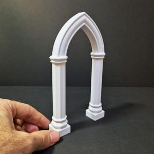 May include: A white, 3D-printed Gothic archway. The arch features two vertical columns with decorative bases and capitals, supporting a pointed arch. The design is a miniature architectural element, suitable for display or crafting projects.