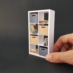 Peut inclure: Étagère blanche à six compartiments. Chaque compartiment contient une boîte de rangement. Les deux compartiments du haut contiennent des livres. Le compartiment du bas contient une pile de livres. Les boîtes de rangement sont grises et beiges.