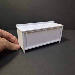 May include: A small, white cabinet with sliding doors and short legs. The cabinet has a rectangular shape with a flat top and a textured surface. The doors appear to be made of a translucent material. The cabinet is designed for miniature use.