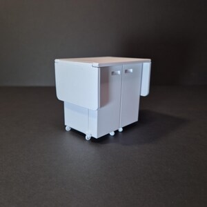 May include: A white, miniature kitchen cart with a drop-leaf table and two cabinet doors. The cart has small wheels for mobility. The drop-leaf table is folded down on one side and up on the other side. The cart is on a dark surface.
