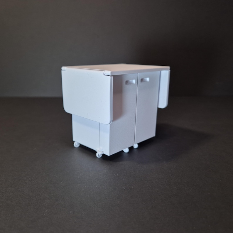 May include: A white, miniature kitchen cart with a drop-leaf table and two cabinet doors. The cart has small wheels for mobility. The drop-leaf table is folded down on one side and up on the other side. The cart is on a dark surface.