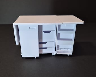 May include: A white miniature craft table with a fold-out extension. It features a cabinet door, three drawers, and open shelving. The table is on wheels and appears to be made of plastic. The table is designed for crafting and sewing.