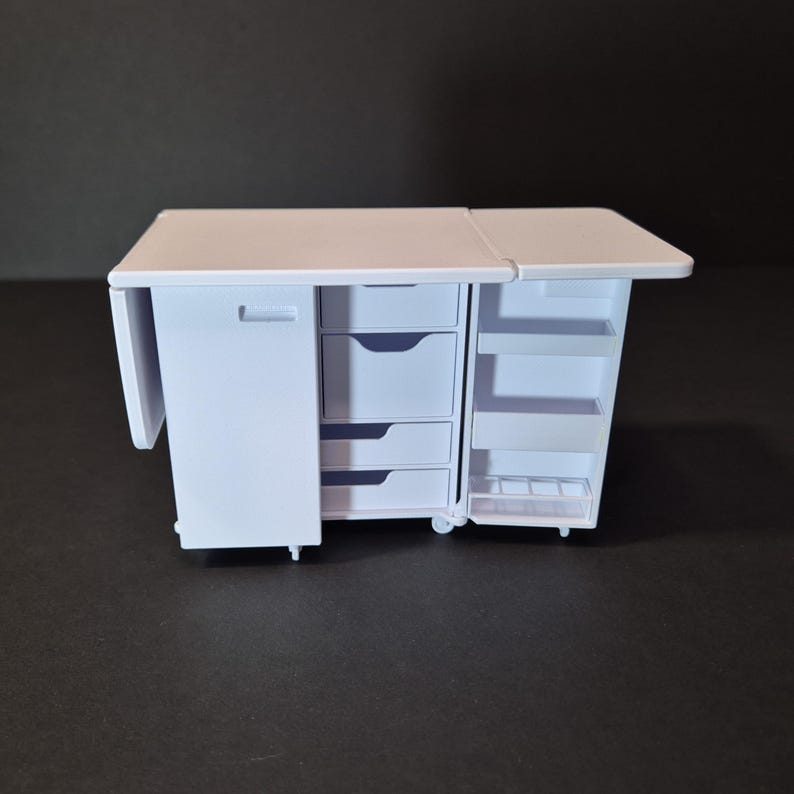 May include: A white miniature craft table with a fold-out extension. It features a cabinet door, three drawers, and open shelving. The table is on wheels and appears to be made of plastic. The table is designed for crafting and sewing.
