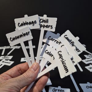 May include: White garden markers with black text, labeled with various vegetables including "Cabbage", "Chili Peppers", and "Cucumber". The markers are held in a hand against a dark background.