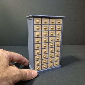 Può includere: Un mobile in miniatura con numerosi piccoli cassetti. Il mobile è dipinto in un grigio-blu tenue, con un fronte in legno chiaro e maniglie dorate. Il mobile è tenuto da una mano.