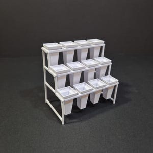 Może przedstawiać: Biały, wielopoziomowy stojak na rośliny z dwunastoma kwadratowymi donicami. Stojak ma prostokątną ramę z trzema poziomami, z których każdy mieści cztery donice. Donice są kwadratowe u góry i zwężają się ku dołowi. Stojak jest metalowy.
