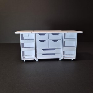 May include: A white miniature craft station with multiple drawers and shelves. The station has a fold-out top and doors that open to reveal storage compartments. The unit is on small wheels, suggesting portability. The overall design is clean and functional.