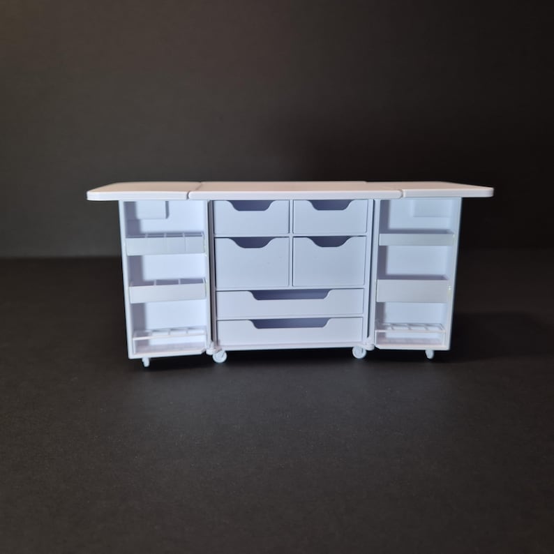 May include: A white miniature craft station with multiple drawers and shelves. The station has a fold-out top and doors that open to reveal storage compartments. The unit is on small wheels, suggesting portability. The overall design is clean and functional.