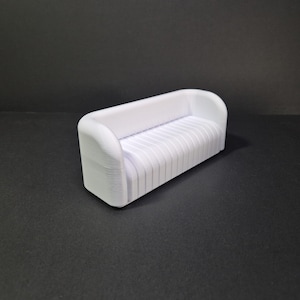 Könnte beinhalten: Ein kleines, weißes Sofa im modernen Stil mit geschwungenem Rücken und Armlehnen. Der Sitz hat vertikale Rippendetails. Das Miniaturmöbelstück steht vor einem dunklen Hintergrund, der seine klaren Linien und sein minimalistisches Design hervorhebt.
