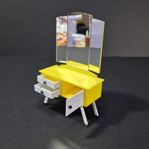 May include: A miniature yellow vanity unit with a tri-fold mirror. The vanity has two drawers and an open cabinet door. The legs are white with silver tips. This is a dollhouse furniture item.