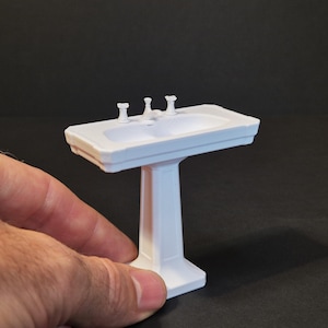 Puede incluir: Un pequeño lavabo de porcelana blanca con dos grifos y una base de pedestal. El lavabo tiene una cubeta rectangular y está diseñado con detalles clásicos. La imagen muestra el lavabo sostenido por una mano sobre un fondo oscuro.