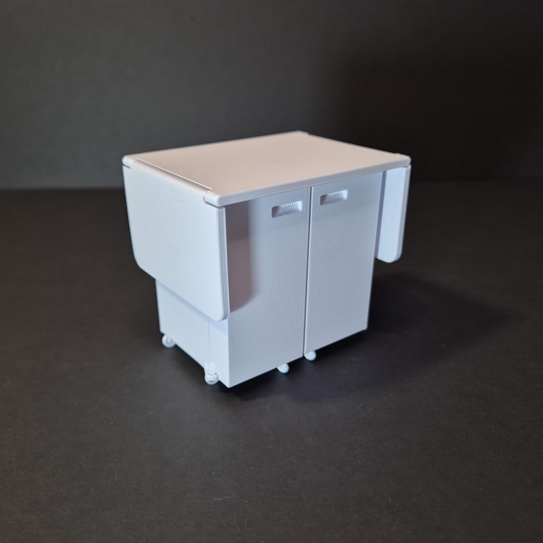 May include: A small, white, miniature cabinet with two doors and a fold-out side panel. The cabinet sits on small wheels and has a rectangular top. The background is a dark gray.