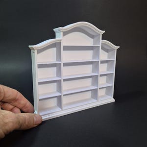 Puede incluir: Una estantería en miniatura de color blanco con una sección central de cuatro niveles y dos secciones laterales, cada una con cuatro estantes más pequeños. La unidad presenta una moldura superior arqueada decorativa y una base plana.