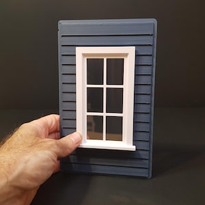 Op de afbeelding: Een wit raamkozijn met zes ruiten glas is in een blauwe gevelbekleding van hout geschroefd. Het raam is een miniatuurmodel, waarschijnlijk voor een poppenhuis of een modelhuis.