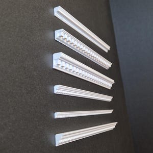 Può includere: Diverse modanature decorative bianche sono disposte diagonalmente su uno sfondo grigio scuro. La modanatura presenta vari disegni dettagliati, tra cui bordi perlati e decorati. I pezzi variano per dimensioni e stile, mostrando elementi architettonici.
