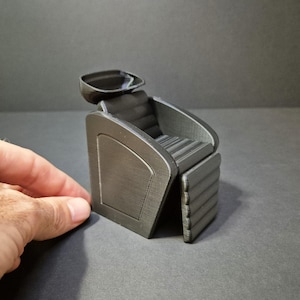 Peut inclure: Une miniature de fauteuil de salon gris foncé avec un appui-tête et des accoudoirs. La chaise a un siège et un dossier côtelés, et un design incurvé. La chaise est fabriquée dans un matériau imprimé en 3D et est posée sur un fond sombre.