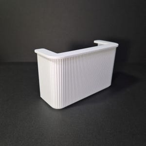 May include: A small, white bar with a ribbed front and a curved countertop. The bar has a contemporary design and is likely for a doll's house or model display. The bar is made of a solid, opaque material.