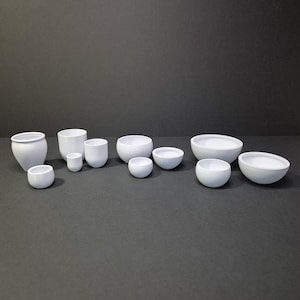 Pode incluir: Uma coleção de tigelas e xícaras de cerâmica branca de vários tamanhos. O sortimento inclui tigelas pequenas e esféricas, xícaras cilíndricas e tigelas maiores e abertas, todas dispostas sobre uma superfície cinza escuro. Os objetos são uniformemente brancos, com superfícies lisas.