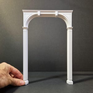 May include: A white miniature archway with a rounded top and fluted columns. The arch is held by a hand, showing its size and detail. The design features classical elements, suitable for model building or decorative purposes.