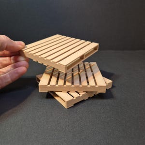 May include: Three small, light-colored wooden pallet coasters. One coaster is being held up, while the other two are stacked on top of each other. The coasters have a slatted design and are made of a light-colored wood.