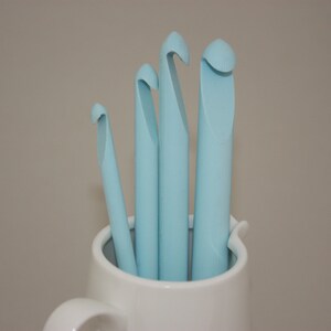 May include: Four light blue crochet hooks of different sizes are standing upright in a white ceramic mug.