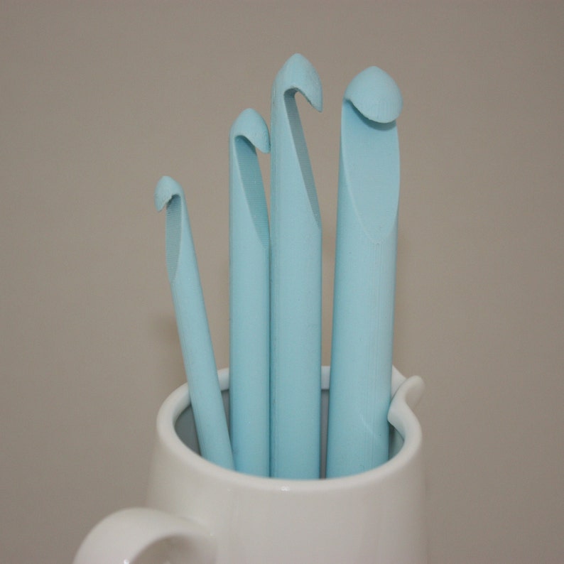 May include: Four light blue crochet hooks of different sizes are standing upright in a white ceramic mug.