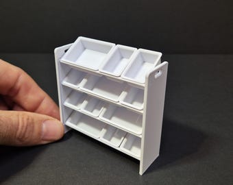 May include: A small, white, multi-tiered shelf unit with several rectangular compartments. The unit has a simple design with a flat base and a series of shelves. The top shelf has three larger compartments, while the lower shelves have smaller ones.