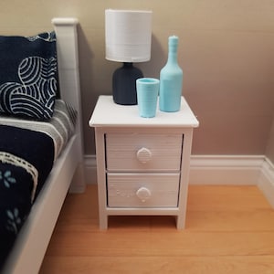 May include: A white nightstand with two drawers and a white lamp with a white shade. There is a blue glass bottle and a blue glass cup on the nightstand.