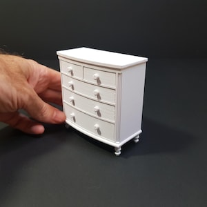 May include: A small, white chest of drawers with five drawers and a curved front. The miniature furniture piece has round knobs and short, rounded legs. The chest of drawers is a miniature replica of a classic design.