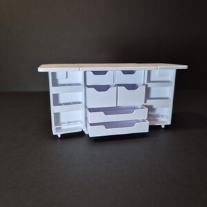 May include: A miniature white cabinet with multiple drawers and shelves. The cabinet has a rectangular shape with a flat top and is designed to open on both sides. The drawers are of varying sizes, and the shelves are open.