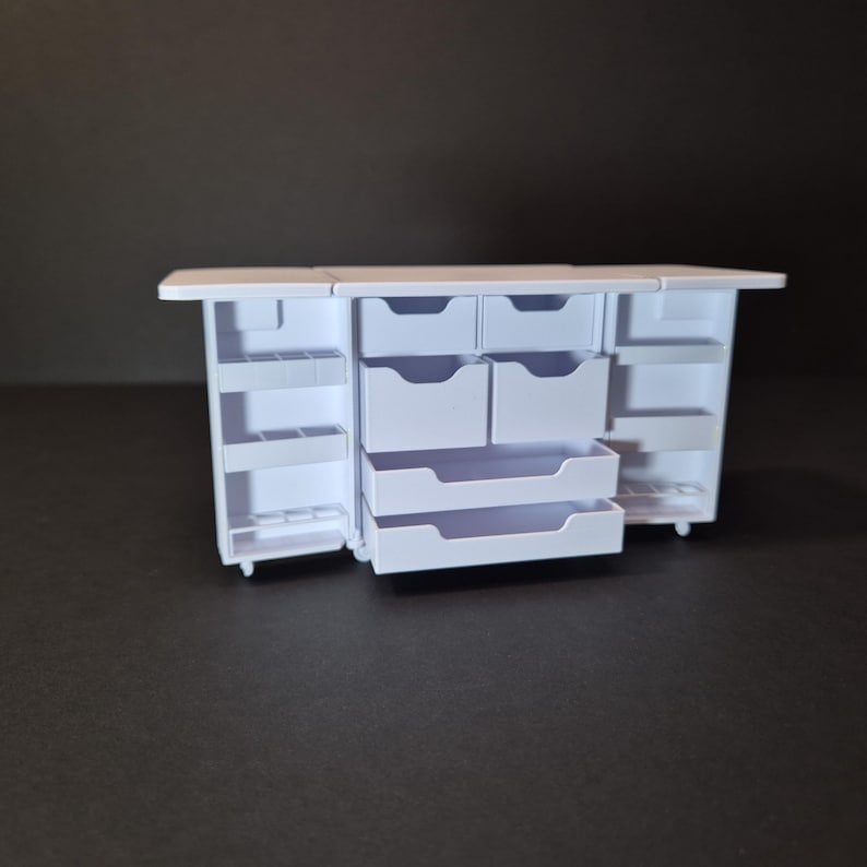 May include: A miniature white cabinet with multiple drawers and shelves. The cabinet has a rectangular shape with a flat top and is designed to open on both sides. The drawers are of varying sizes, and the shelves are open.