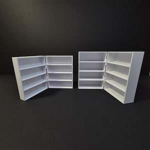 May include: Four small, white, hinged bookshelves are arranged against a black background. Each bookshelf has multiple shelves. Two of the bookshelves are open, revealing the interior shelves, while the other two are closed.