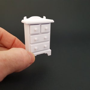 May include: White miniature dresser with four drawers and a simple design. The dresser has a slightly rounded top and sits on four small feet.