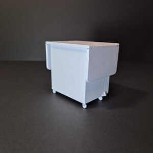 May include: A small, white, miniature cabinet with a drop-leaf table design. The cabinet has a rectangular shape and sits on four small wheels. The drop-leaf is folded down, and the cabinet is painted in a solid white color.