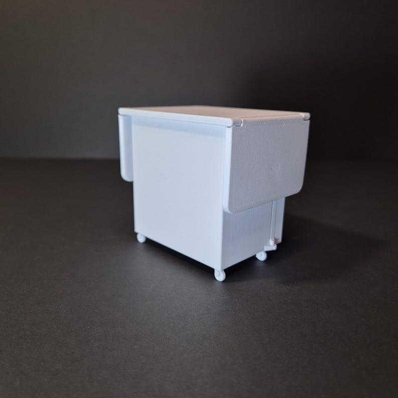 May include: A small, white, miniature cabinet with a drop-leaf table design. The cabinet has a rectangular shape and sits on four small wheels. The drop-leaf is folded down, and the cabinet is painted in a solid white color.
