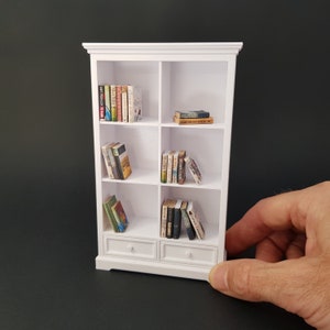 May include: White miniature bookshelf with six shelves and two drawers. The shelves are filled with miniature books.