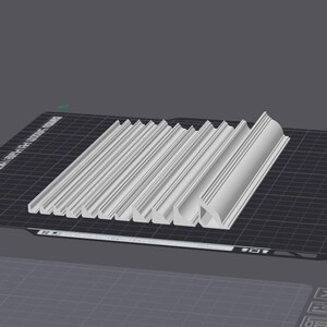 May include: A close-up of a set of 12 white plastic moldings, each with a slightly different shape and size. The moldings are arranged in a row on a black grid surface.