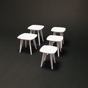 May include: A collection of six small, white, square-topped tables with angled legs. The tables are arranged on a dark background. Each table has a simple, minimalist design, suitable for various decorative or functional purposes.
