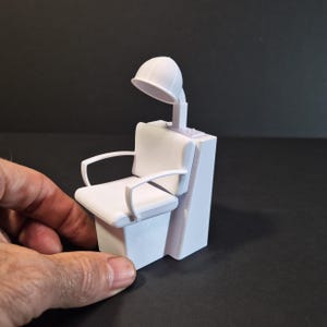 May include: A small white salon chair with a hooded hair dryer. The chair features armrests and a cushioned seat and back. The hair dryer is connected to the chair's back. The object is held by a hand.