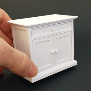 May include: White wooden cabinet with a single drawer and two doors. The cabinet has a simple design with a slightly raised base and a decorative trim around the top.