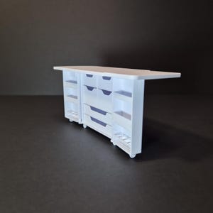 May include: A white miniature kitchen island with drawers, shelves, and a countertop. The island has a rectangular shape and is set on small wheels. The drawers and shelves are visible on the sides and front of the island.