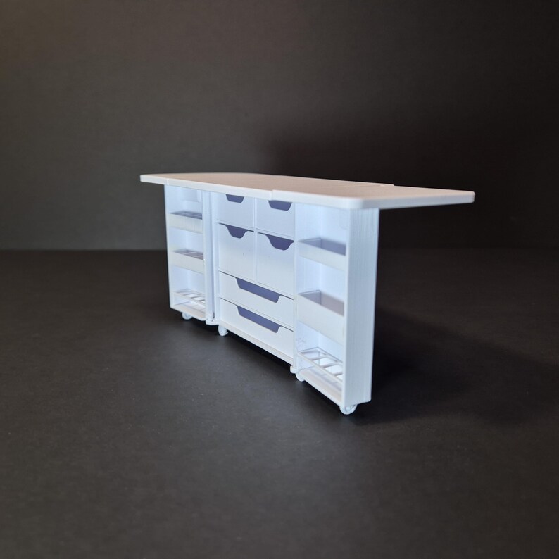 May include: A white miniature kitchen island with drawers, shelves, and a countertop. The island has a rectangular shape and is set on small wheels. The drawers and shelves are visible on the sides and front of the island.
