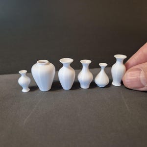 Könnte beinhalten: Satz von sieben weißen Keramikvasen in verschiedenen Größen und Formen. Die Vasen sind in einer Reihe auf einer grauen Oberfläche angeordnet.